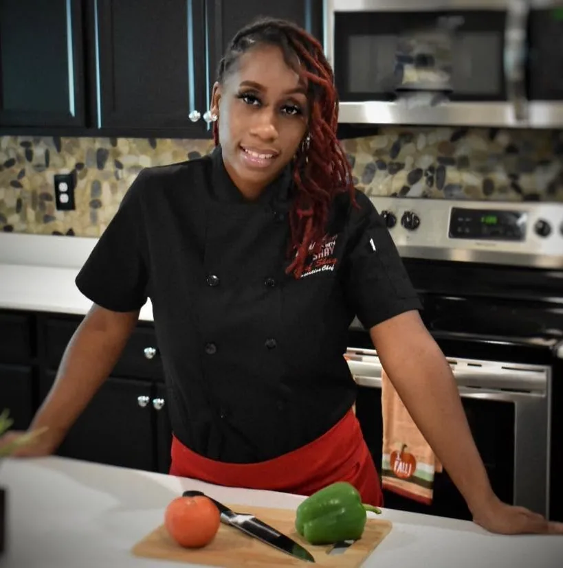 Chef Shay in a black Mixson With Shay chef coat and red apron at a kitchen counter